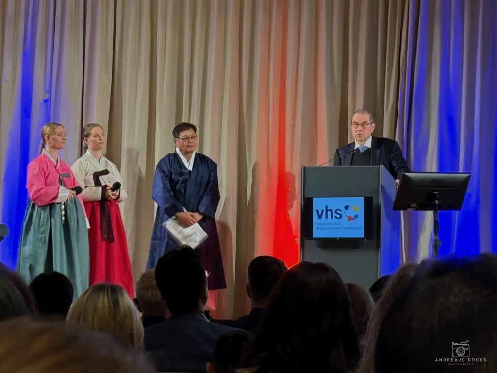 Die Moderatorinnen Jil und Gina Kany, Jae Sun Byun, Vorsitzender der Koreaner im Saarland und Prof. Dr. Rolf Hempelmann, Vorsitzender der Deutsch-Koreanischen Gesellschaft RV Saarland am Rednerpult